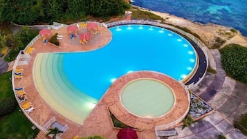 An aerial view of a resort with a large circular pool surrounded by lounge chairs and umbrellas, with a beach and ocean in the background.