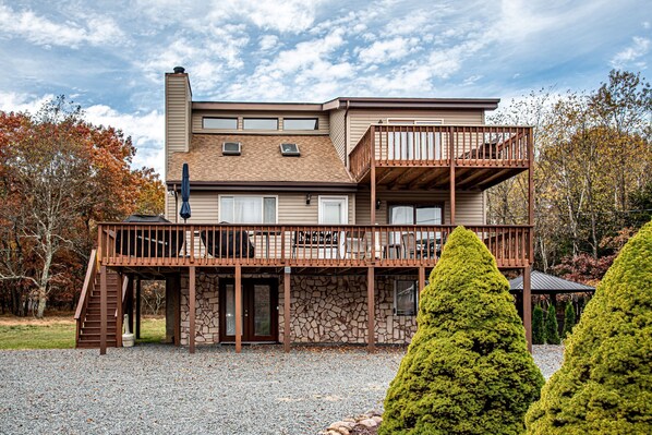 A two-story house with a balcony, stone foundation, and a gravel driveway.