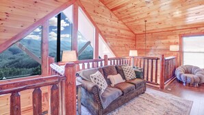 A cozy cabin interior with a wooden ceiling, a large window offering a mountain view, a cushioned sofa, and a wooden railing.