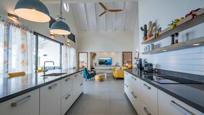 A modern kitchen with white cabinets, black countertops, and a central island. There is a ceiling fan, a television, and a dining area with yellow chairs. - Beautiful villa with breathtaking views and direct access to the sea (Rif St. Marie)