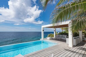 En infinitypool med ett trädäck, vita utemöbler och utsikt över havet.