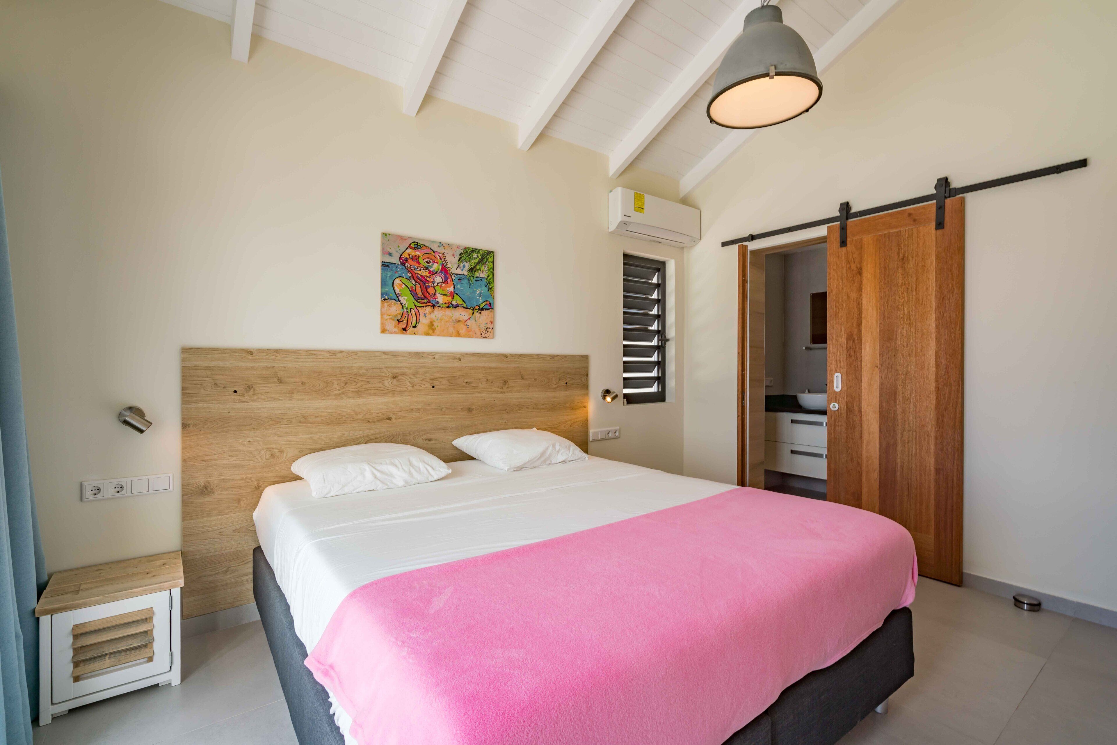 Un dormitorio con cabecera de madera, cama con ropa de cama blanca y una manta rosa, una lámpara de techo y un armario con puerta corredera.