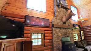 L’intérieur d’une cabane en rondins, avec une cheminée en pierre, des murs en bois, une tête de cerf accrochée au mur, un téléviseur et un canapé.