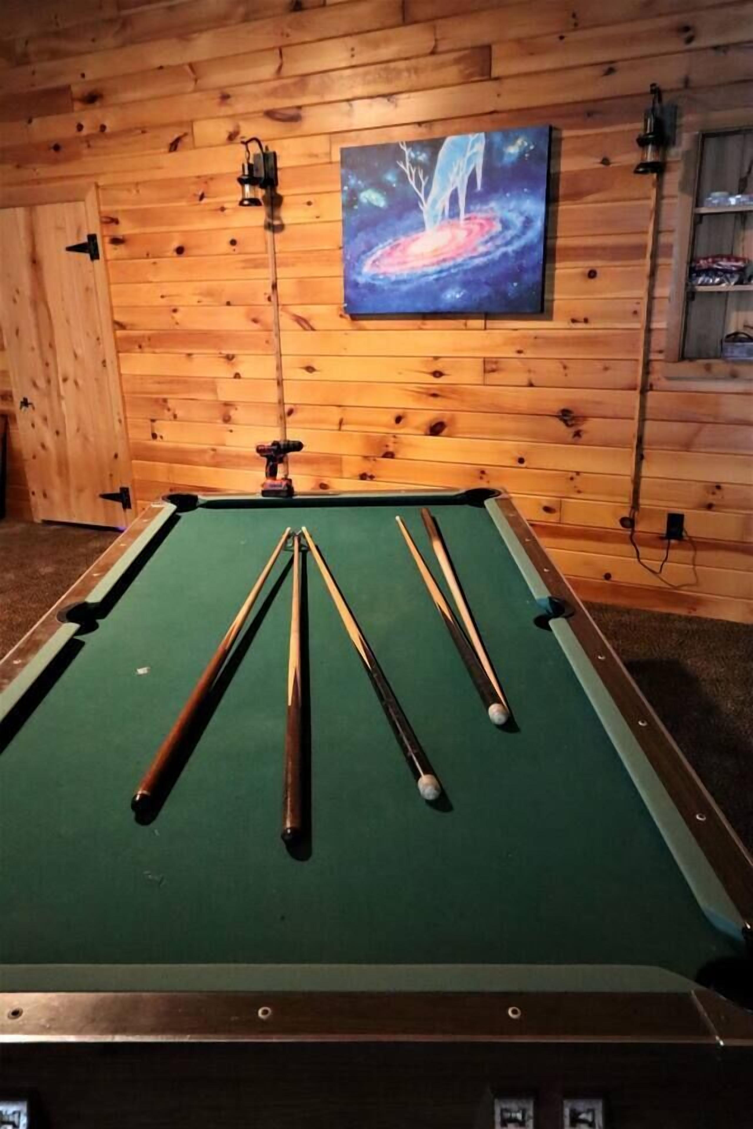 Una mesa de billar con tacos y una bola, frente a una pared de madera con un cuadro.