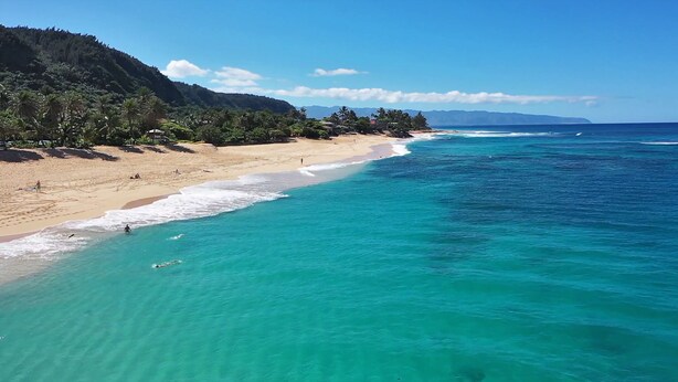 Una playa con agua turquesa cristalina, orilla arenosa y un promontorio boscoso.