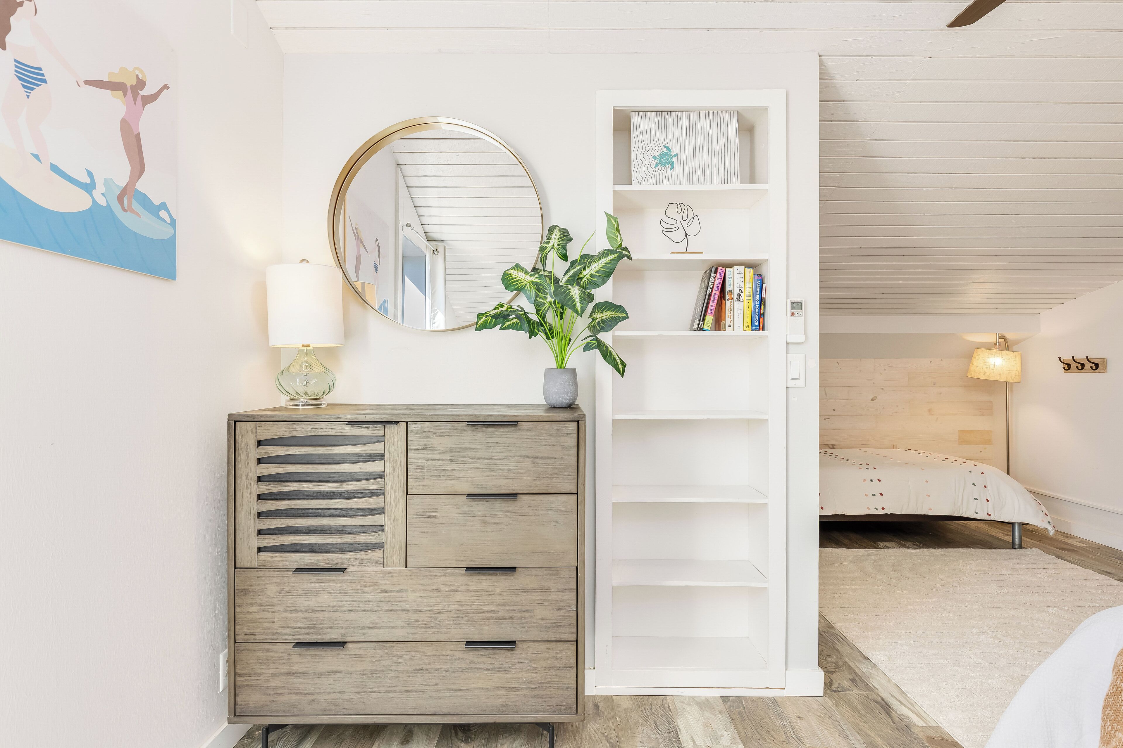 A bedroom with a bed, a bedside table with a lamp, a dresser, a mirror, and a plant.