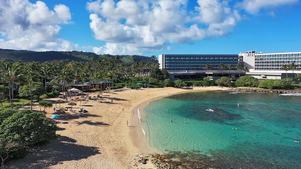 A beach resort with a sandy shore, clear turquoise water, and a large hotel building with multiple floors.