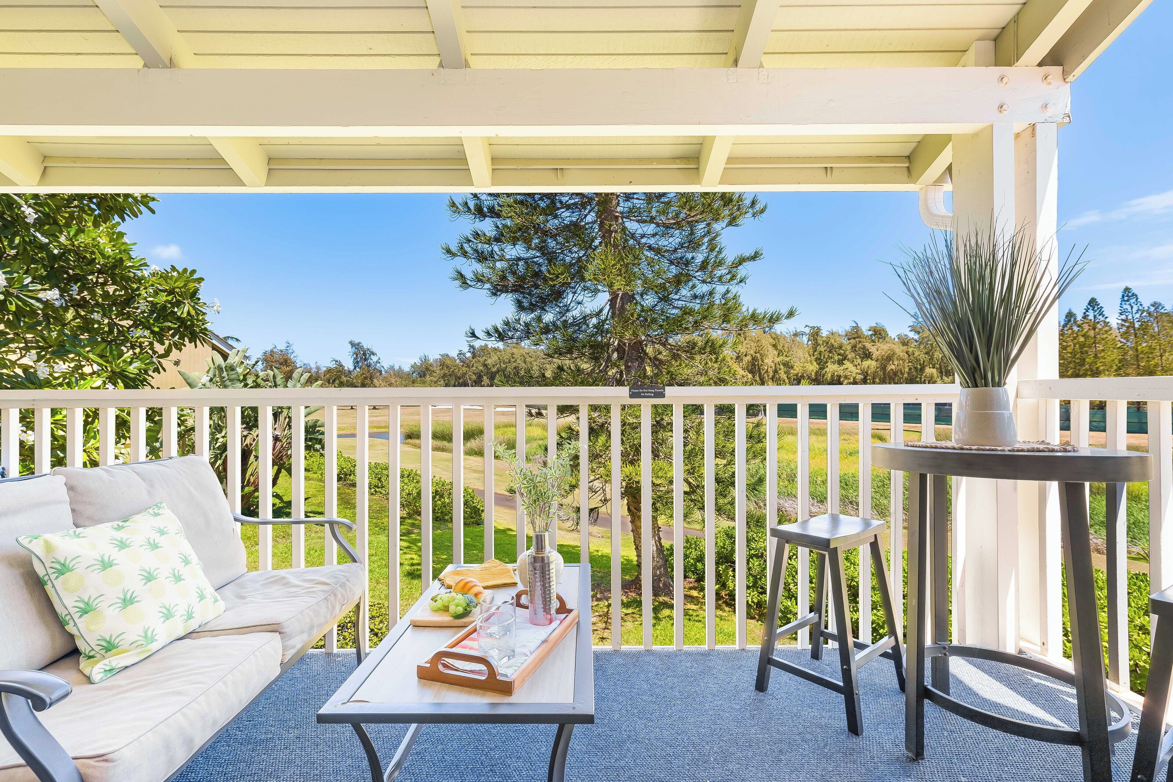 Un patio couvert avec une balustrade blanche, un canapé agrémenté d’un coussin à motifs d’ananas, une table sur laquelle se trouve un plateau de fruits et des verres, et une plante en pot.