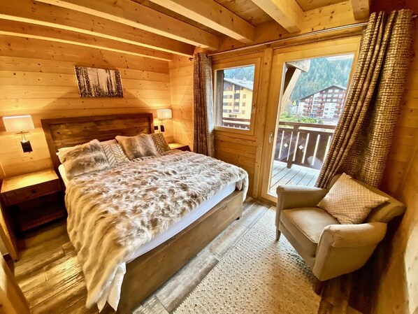 A cozy wooden room with a large bed, a chair, and a balcony with curtains.