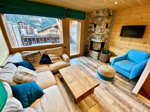 A cozy room with a sofa, a blue armchair, and a wooden coffee table. A stone fireplace is visible, and there is a window with a view of buildings and mountains.