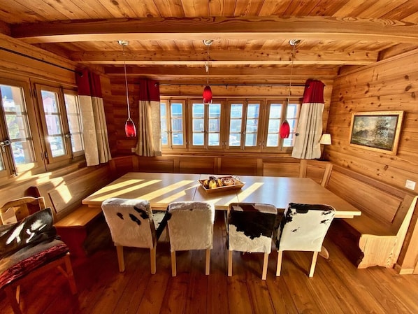 Ein Holzblockhaus-Innenraum mit Esstisch, vier Stühlen mit Kuhfellbezug und einem Fenster mit Vorhängen.