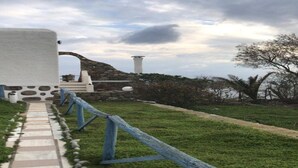 A pathway with a stone border, a grassy area, and a stone structure with a white wall and a blue railing. - Rare and magical panoramic view of Mykonos and the Aegean sea. (Mikonos)