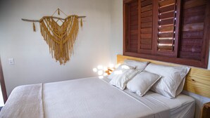 Una cama con ropa blanca, un tapiz de macramé y una ventana con contraventanas de madera.