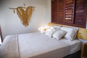 Cama com roupa de cama branca, um pendente de macramé na parede e janela com persianas de madeira.