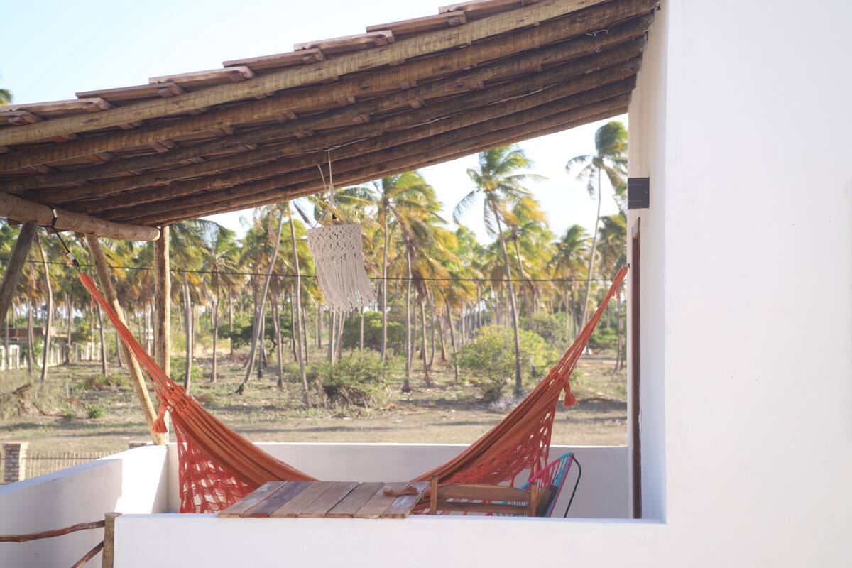 A hammock under a wooden roof, overlooking a beach with palm trees.