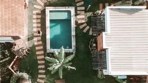 Una piscina rectangular rodeada por un sendero de ladrillo, con una cerca blanca y un pequeño jardín con palmeras.