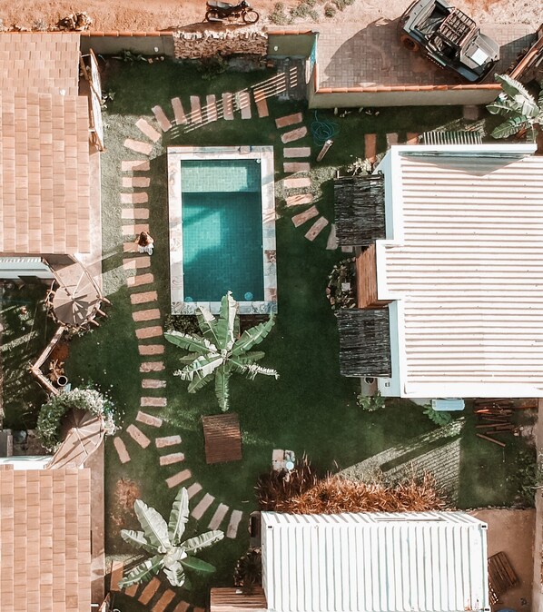 A rectangular swimming pool surrounded by a brick pathway, with a white fence and a small garden area with palm trees. - Zero Meia Um pousada (São Miguel do Gostoso)