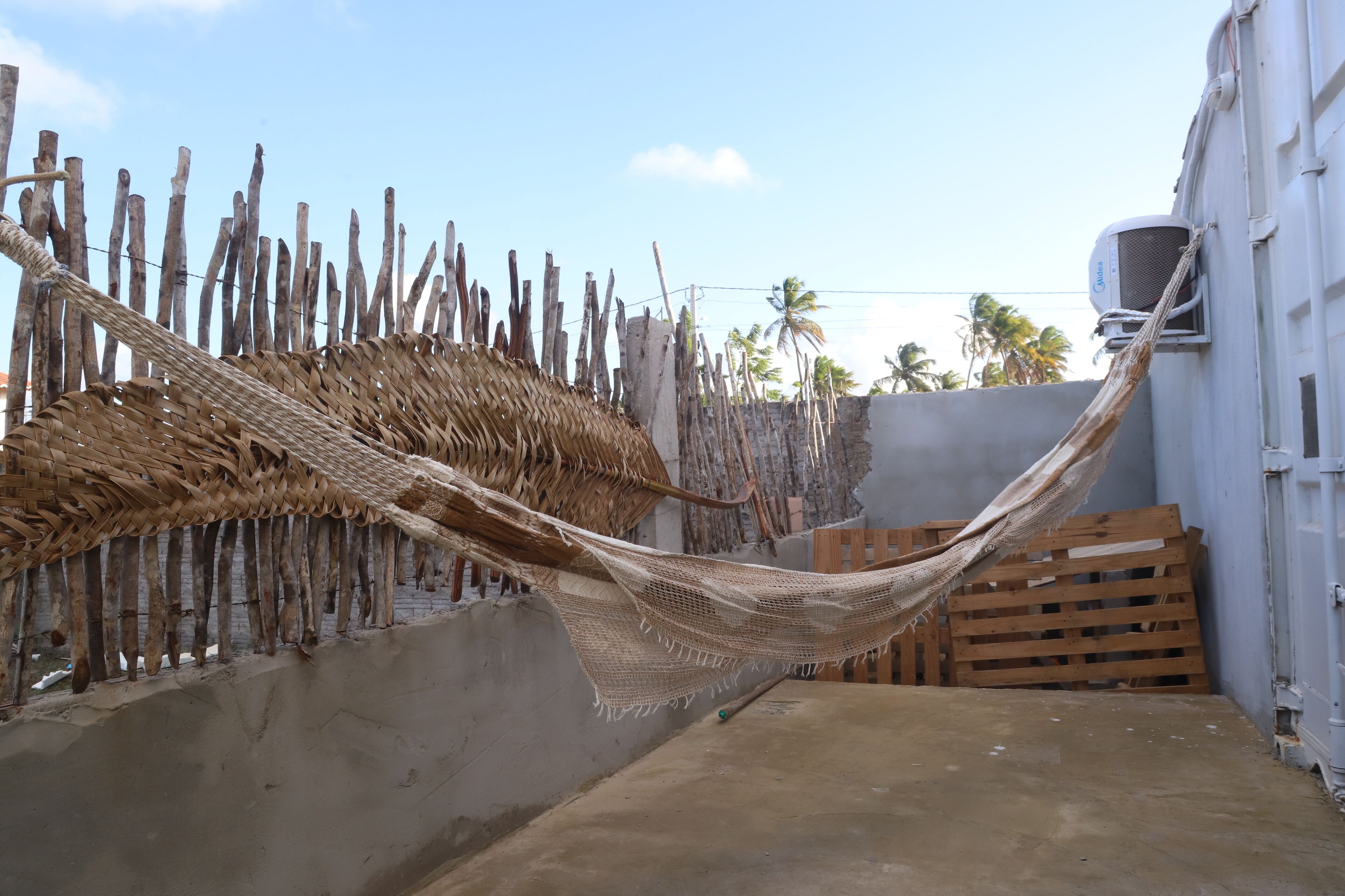 Un hamac est tendu entre un mur de béton et une clôture en bois, avec un palmier et un bâtiment en arrière-plan.