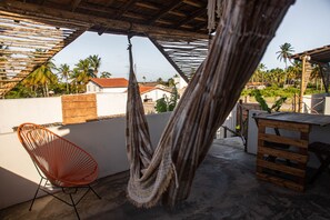 A hammock, a red chair, and a wooden table on a rooftop patio with a view of palm trees and houses. - Zero Meia Um pousada (São Miguel do Gostoso)