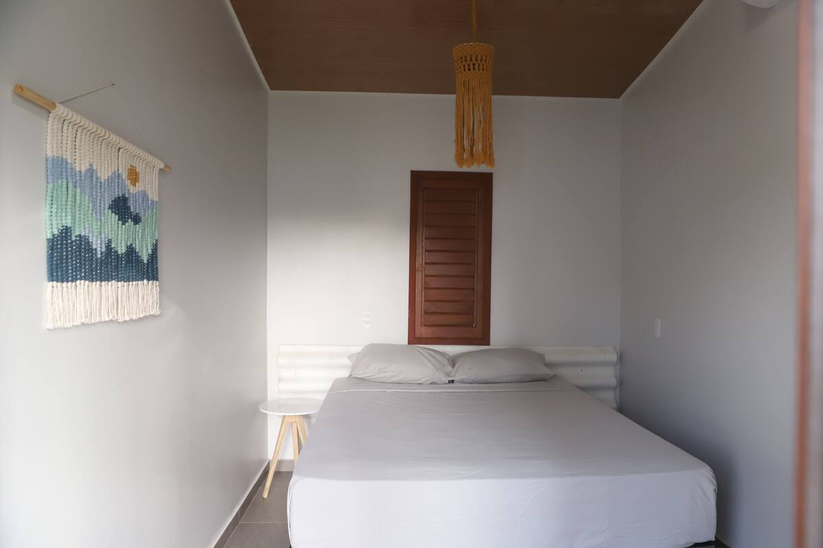 A bedroom with a bed, a wooden door, a hanging light fixture, and a wall-mounted macrame piece.