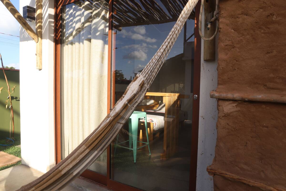 A hammock tied between a window and a wall, a green chair, and a bed with a white and red patterned bedspread.