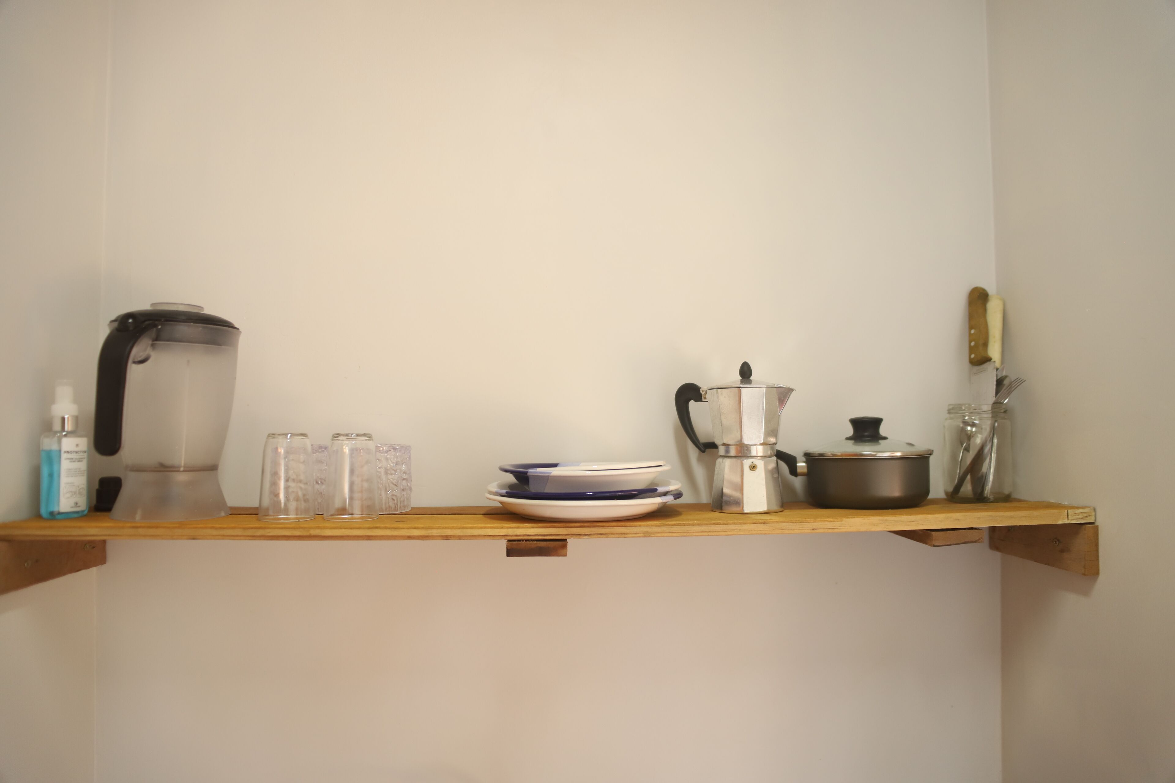 A kitchen shelf with a kettle, coffee pot, plates, cups, and a jar with a spoon.