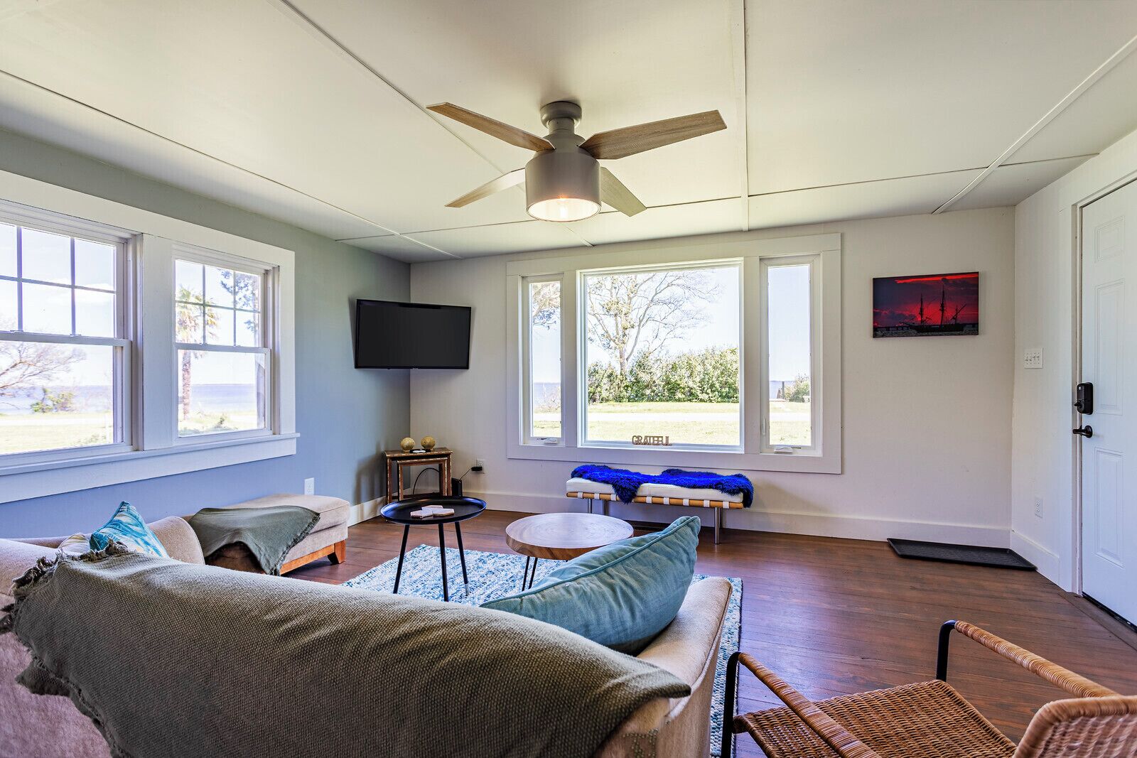A living room with a ceiling fan, large windows, a flat-screen TV, and a bench by the windows.