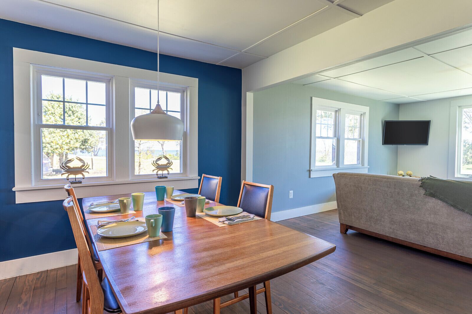 A dining area with a wooden table set for four, surrounded by blue walls and large windows.