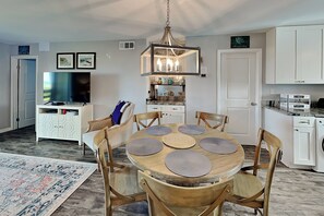 Uma sala de jantar com uma mesa de madeira redonda, seis cadeiras, um candelabro, uma televisão e uma cozinha ao fundo.