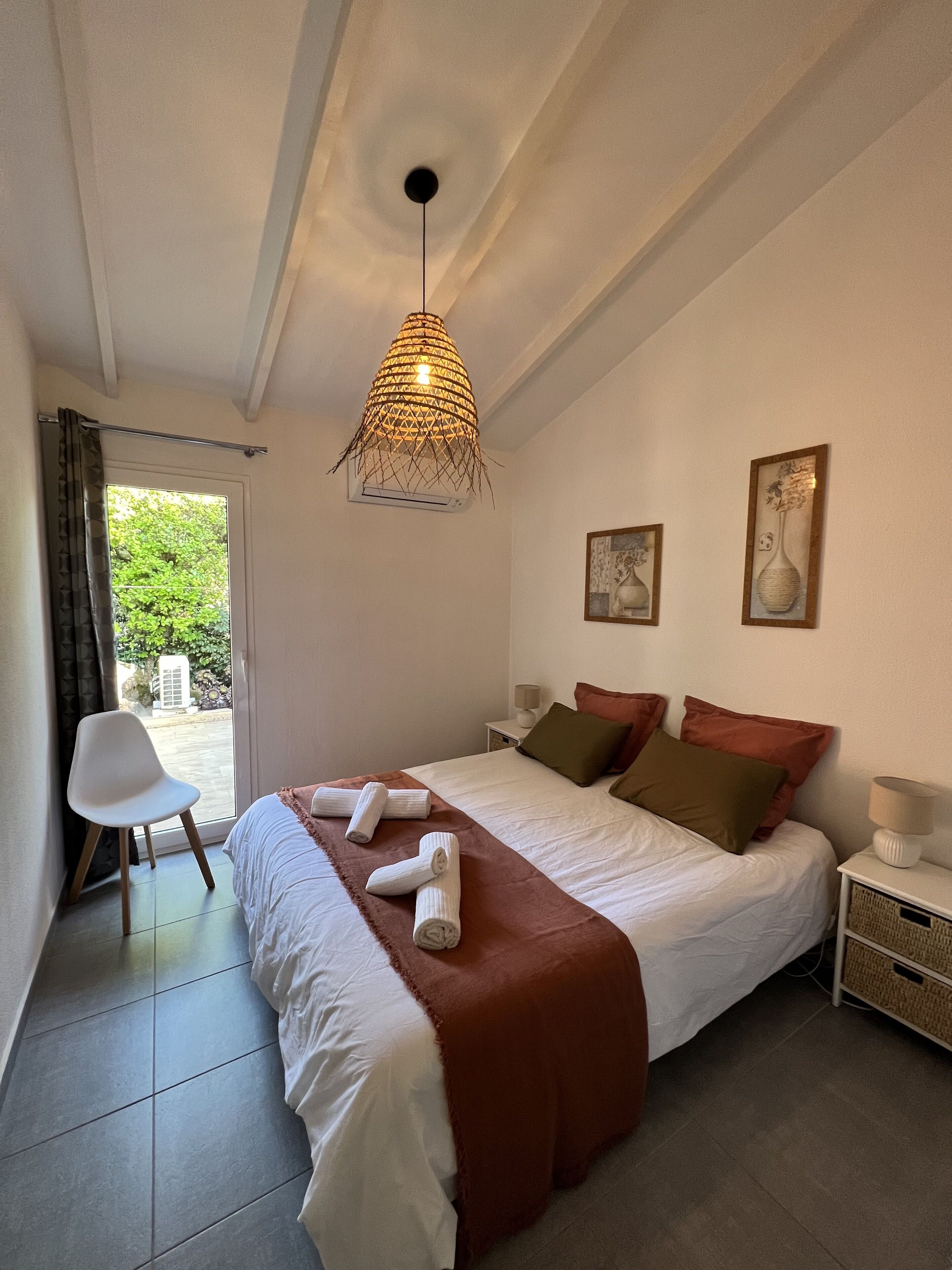 A neatly made bed with a brown blanket, a white chair, and a hanging light fixture.