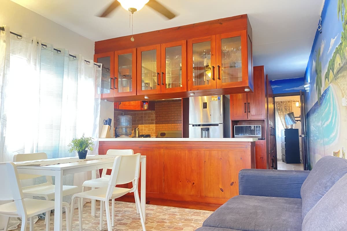A kitchen with wooden cabinets, a refrigerator, and a water dispenser. There is a dining table with chairs and a sofa in the living area.