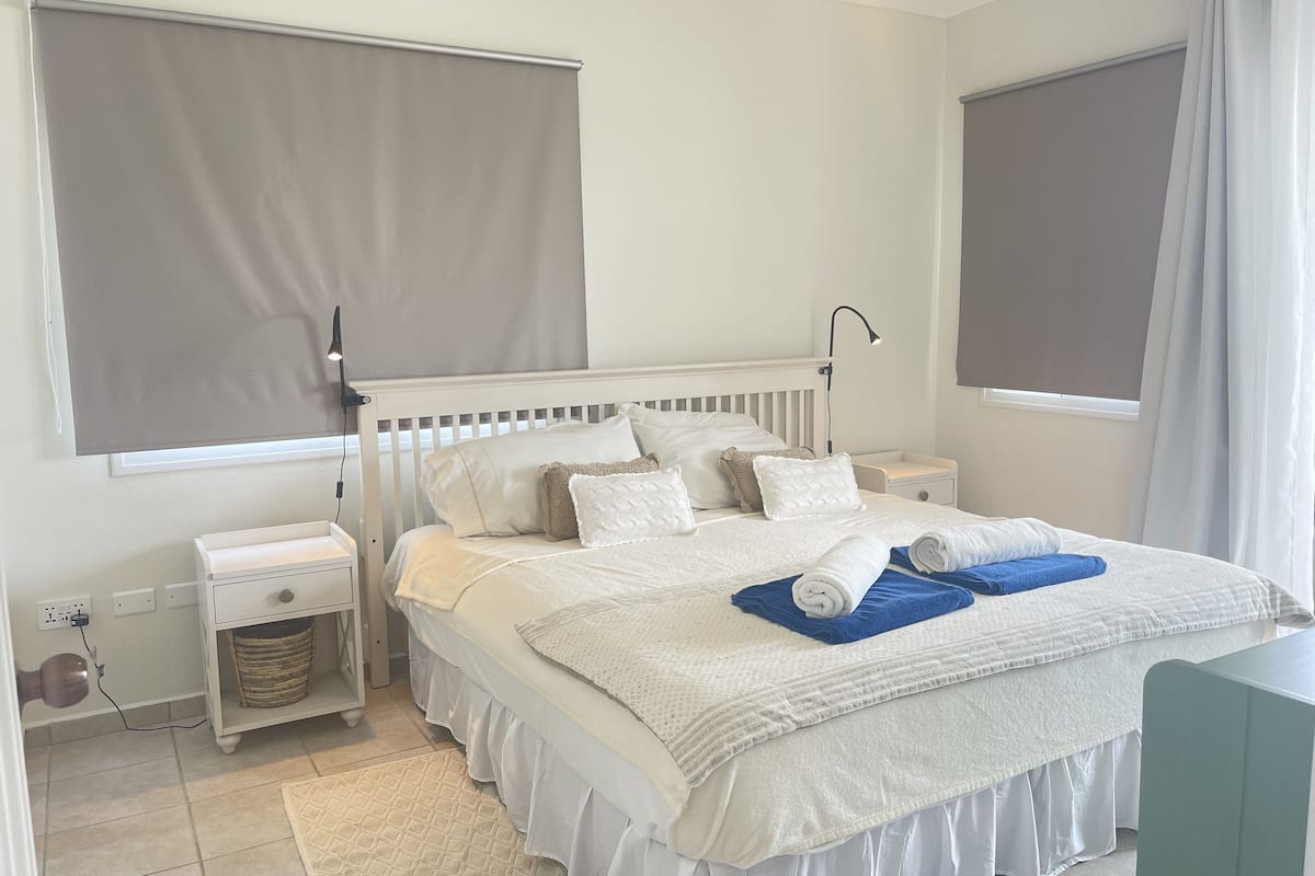 A bedroom with a white bed, a bedside table, a lamp, a waste basket, and a window with a blind.