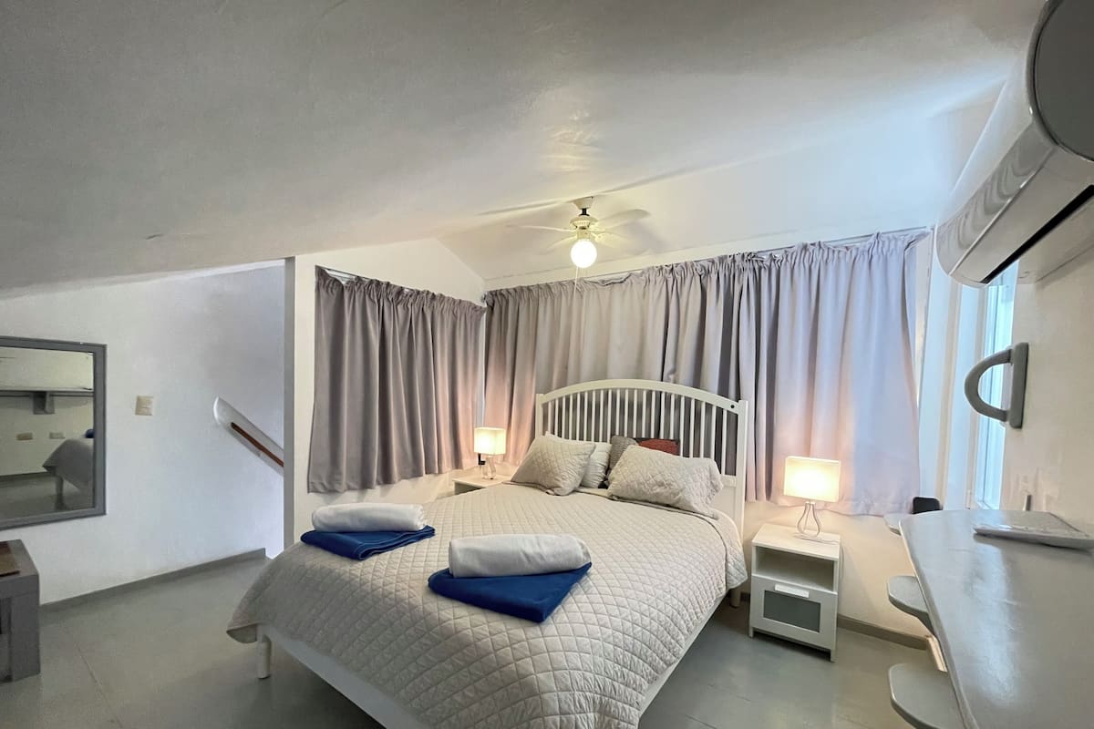 A bedroom with a bed, nightstands, a desk, a ceiling fan, and curtains.