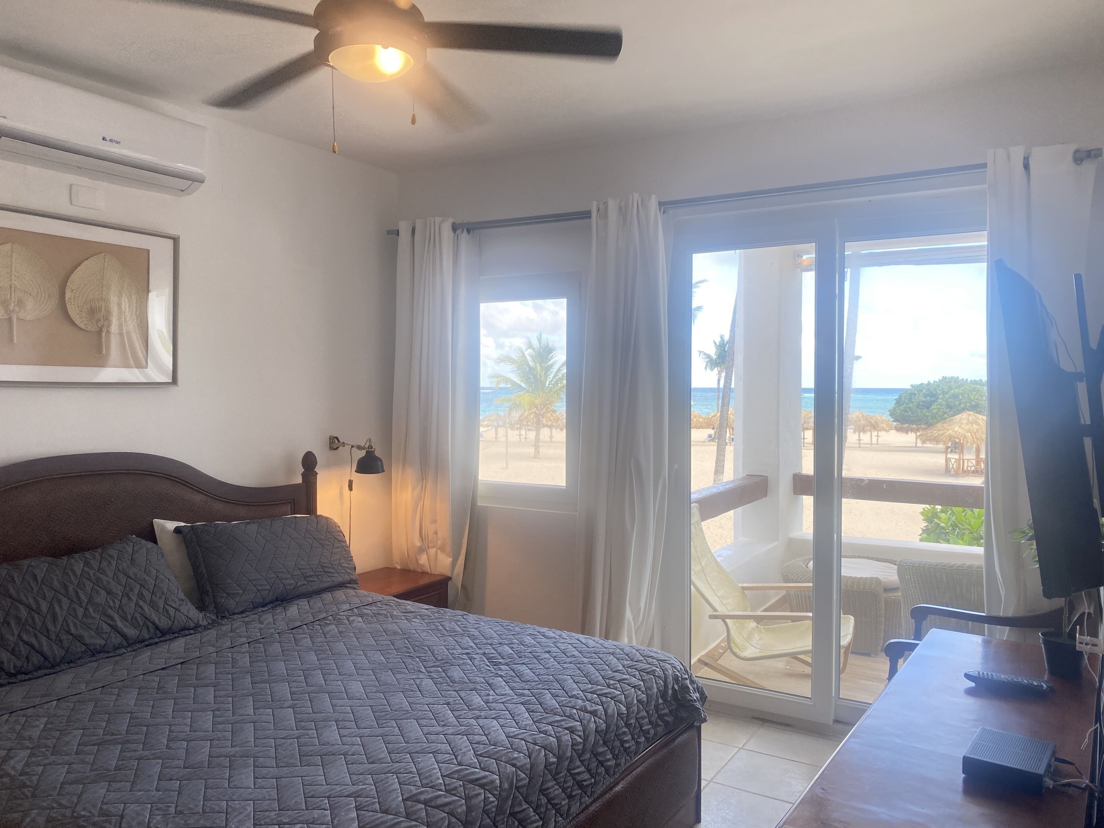 A bedroom with a bed, a ceiling fan, a nightstand, a desk with a computer and a remote control, and a sliding glass door leading to a balcony with a beach view.
