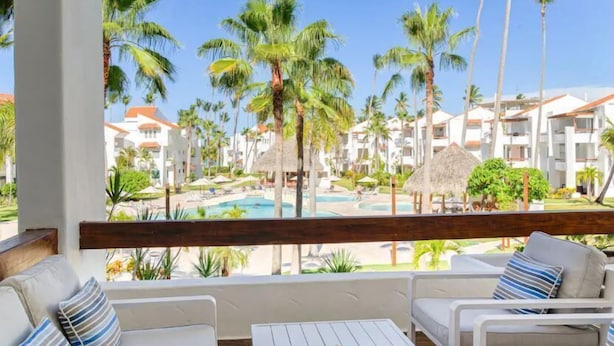 Balcón de un resort con mobiliario blanco, piscina y palmeras.