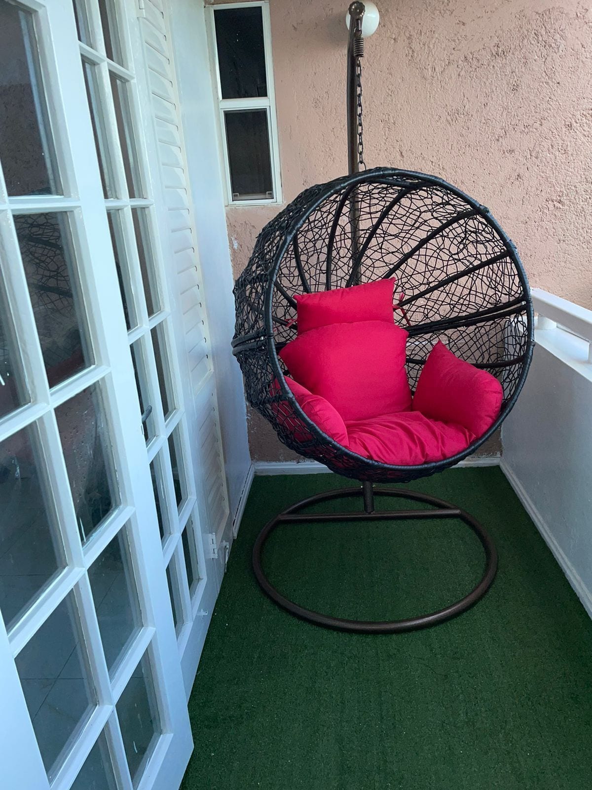 A corner room with a white-framed window, a hanging chair with red cushions, and a green artificial turf floor.