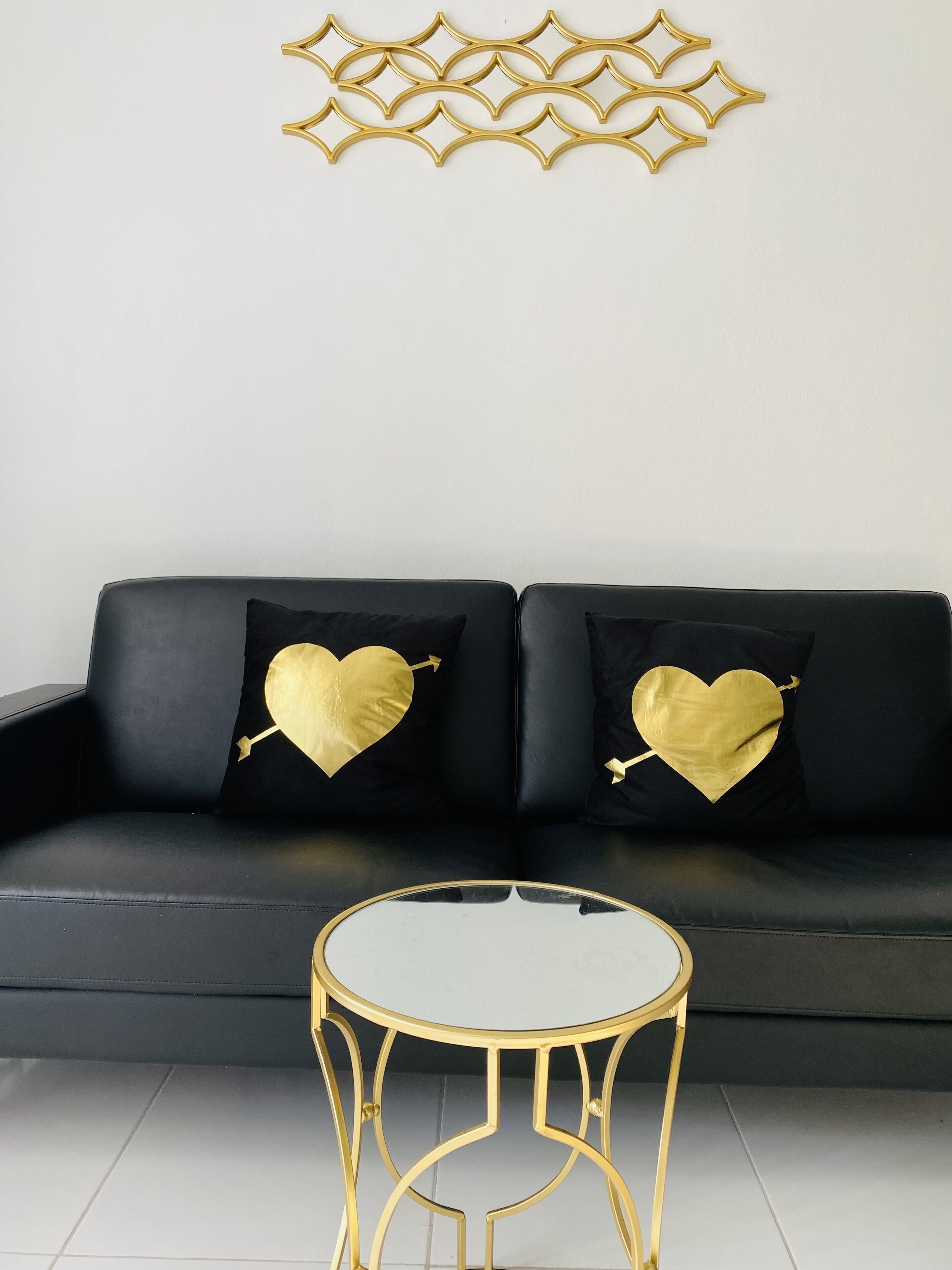 A black leather sofa with two black cushions featuring gold heart designs and arrows. In front of the sofa is a small round table with a gold frame.