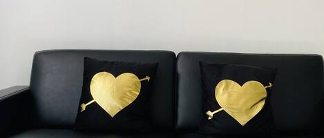 A black leather sofa with two black cushions featuring gold heart designs and arrows. In front of the sofa is a small round table with a gold frame.