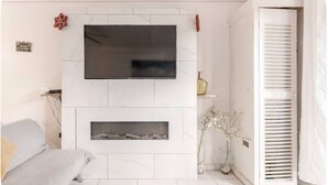 A modern living room with a flat-screen TV mounted on a white tiled wall, a fireplace below, a sofa, a vase with flowers, and a white door with shutters.