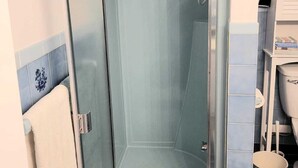 A glass-enclosed shower stall with a white door, a toilet, and a small trash bin.