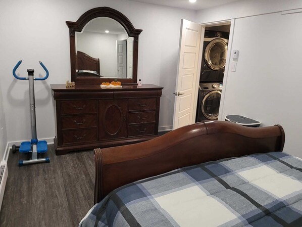 A bedroom with a wooden bed, a dresser, a mirror, a vacuum cleaner, and a washing machine.