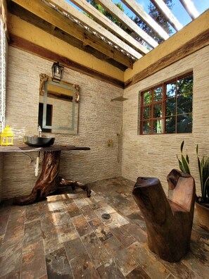 A rustic bathroom with a stone wall, a wooden vanity, a mirror, and a window. - Ecoscape Jamaica - 2 Bedroom Cottage by the river - 30 Minutes to Ocho Rios (St. Ann Parish)