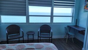 A room with a bed, two wicker chairs, a small table, a desk, and a view of the ocean through large windows.