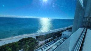 A view of a beach with a clear sky and a bright sun reflecting on the water. There is a building with a balcony overlooking the beach.