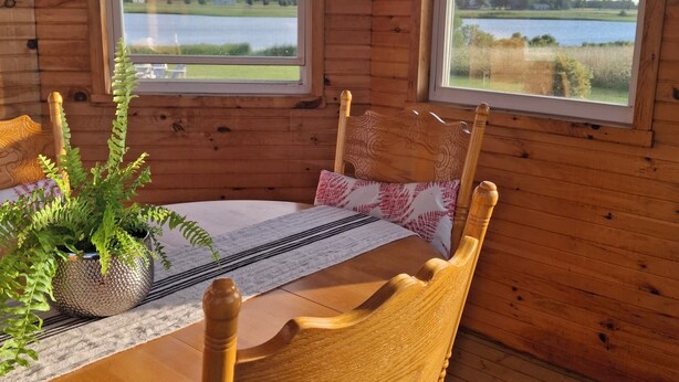 Puinen mökki sisältä, jossa on ruokapöytä, tuoleja ja näkymä järvelle kahden ikkunan kautta.
