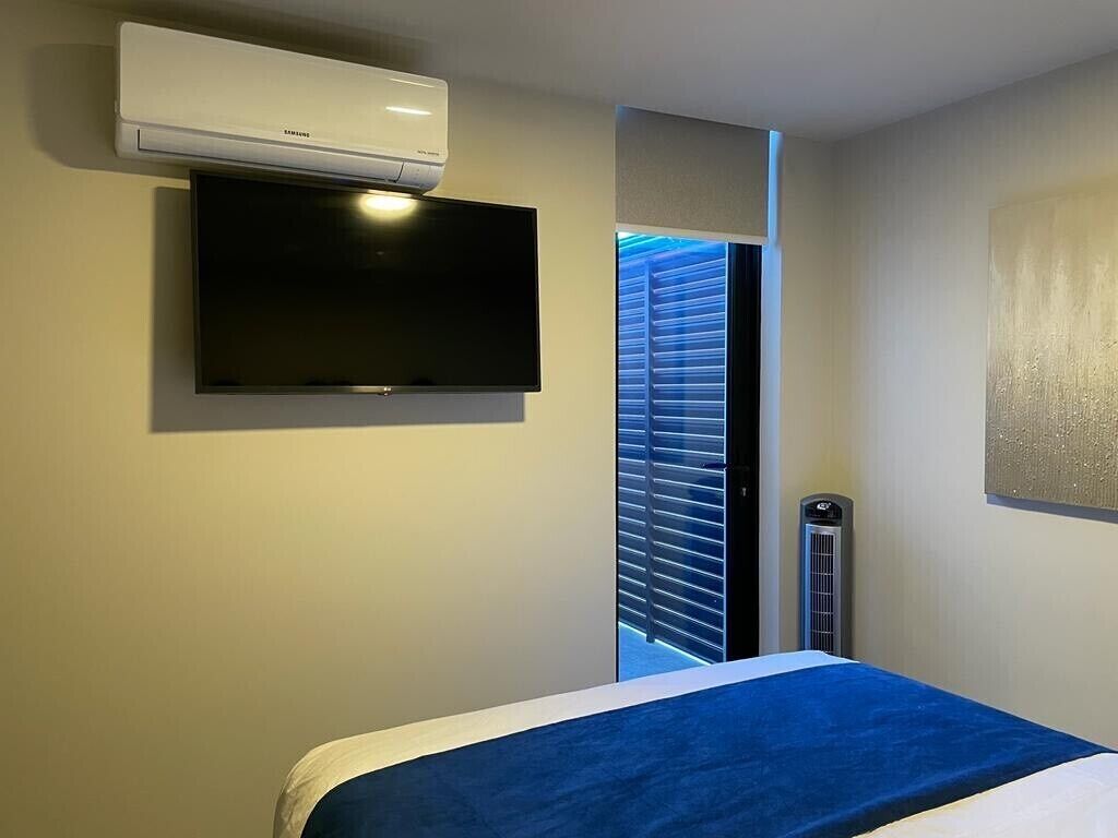Hotellihuone, jossa on sänky, seinälle asennettu taulutelevisio, ikkuna, jossa on sälekaihtimet, ja seinälle asennettu ilmastointilaite.