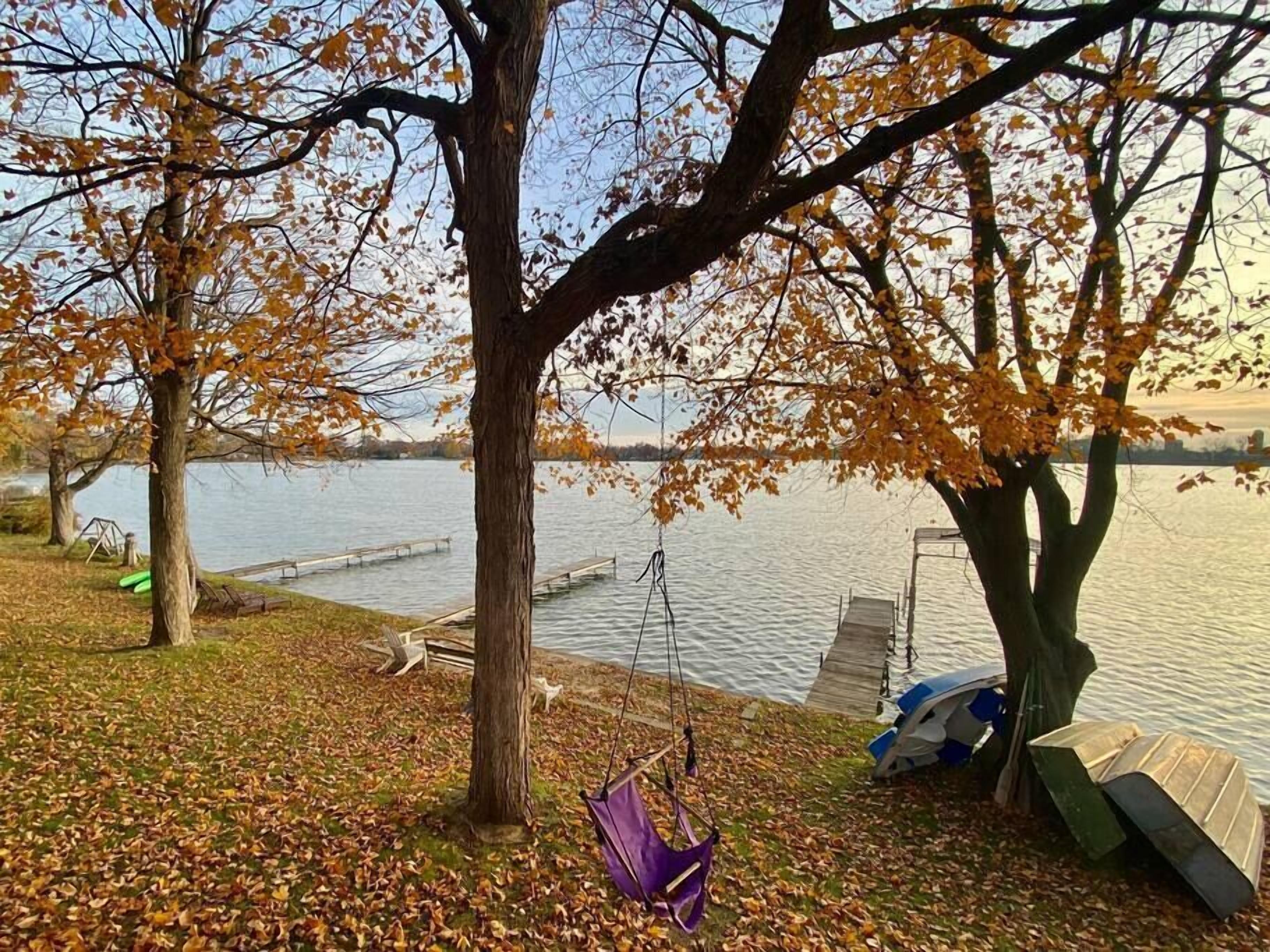 Escena junto al lago con un muelle, una hamaca y un árbol con hojas otoñales.