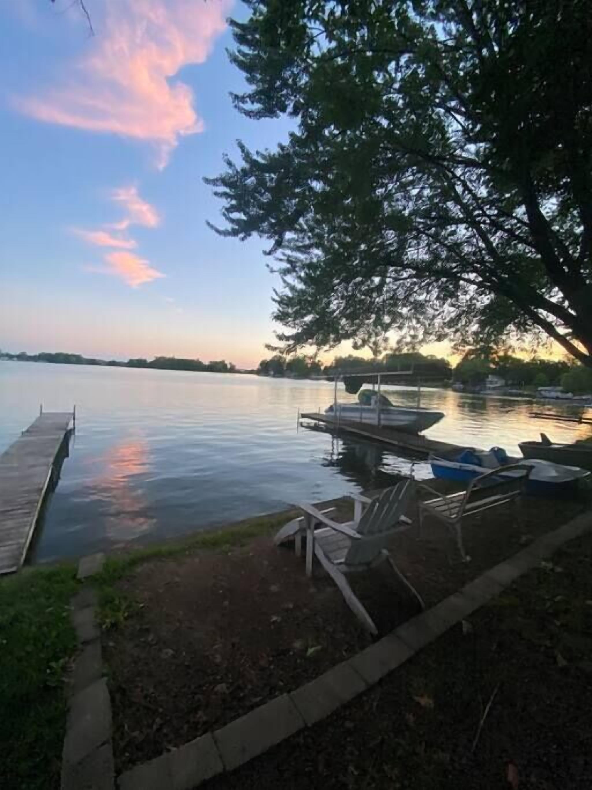 A lakeside dock with boats and chairs, a wooden pier, and a tree providing shade.