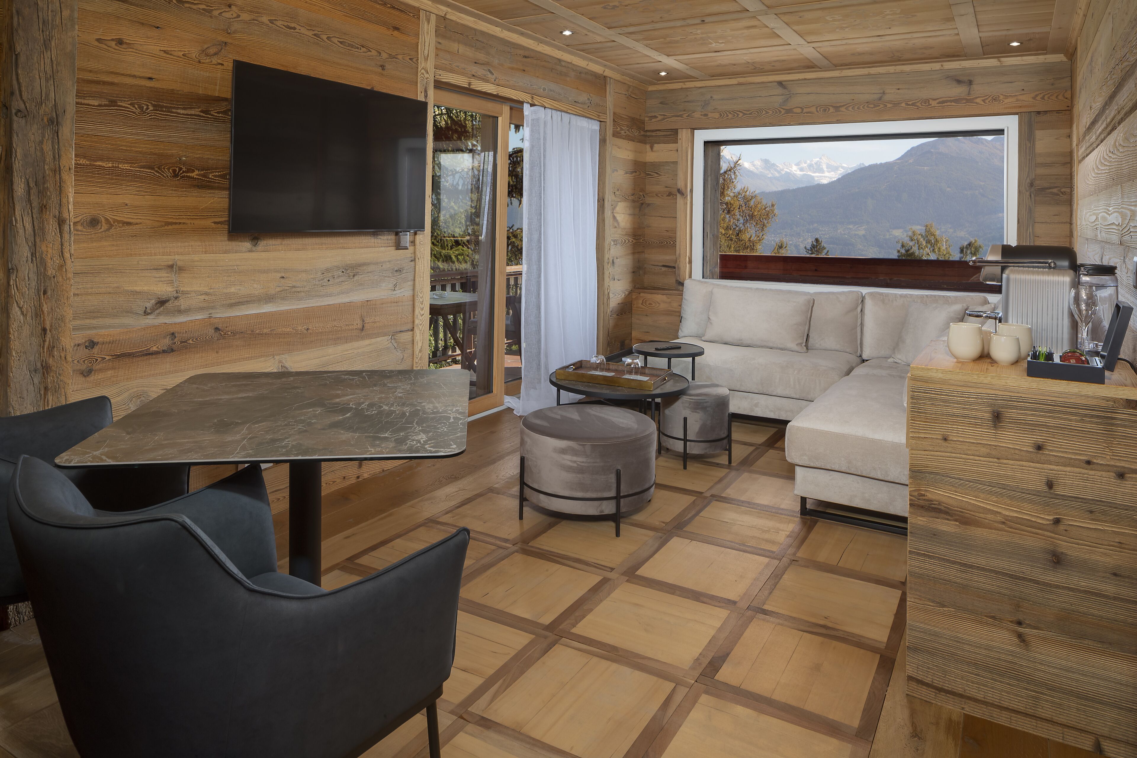 Ein gemütliches Blockhäuschen im Inneren mit Holzausstattung, einer Couch, einem Esstisch mit Stühlen und einem großen Fenster mit Blick auf die Berge.