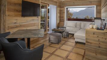 Ein gemütliches Blockhäuschen im Inneren mit Holzausstattung, einer Couch, einem Esstisch mit Stühlen und einem großen Fenster mit Blick auf die Berge.